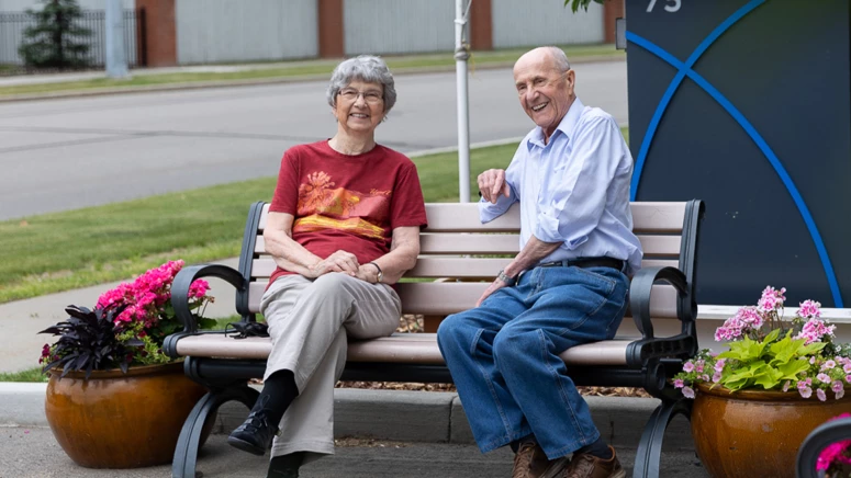 A senior couple sitting on a bench beside potted flowers outside a retirement home